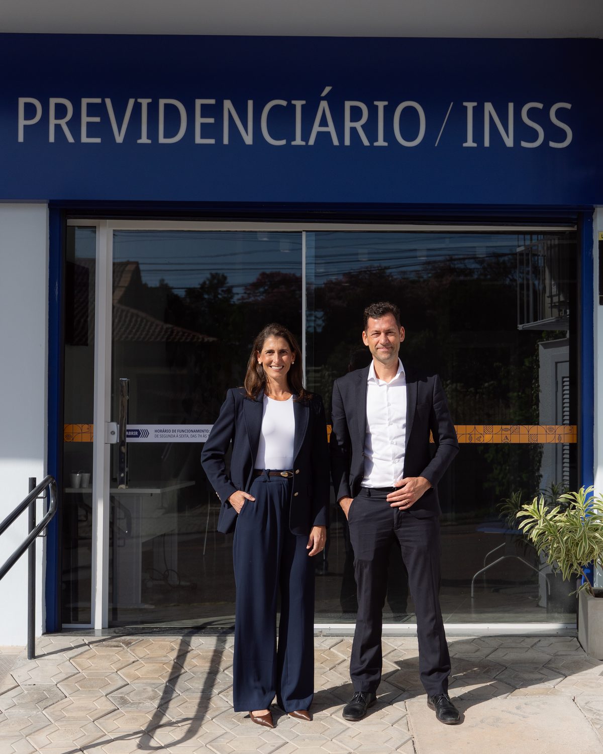 Dra. Fernanda Dutra e Dr. Gustavo Dutra na entrada do Dutra Advogados, sob o letreiro Previdenciário / INSS